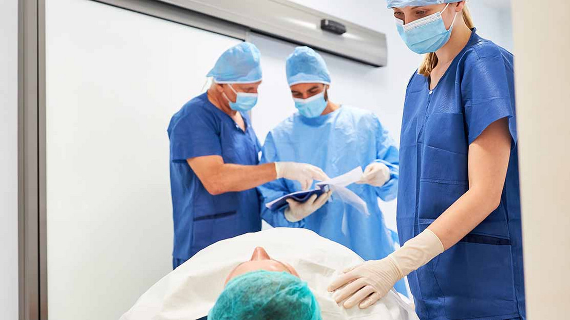 Drei Pfleger oder Ärzte mit blauen Kitteln stehen an einem Patientenbett. 