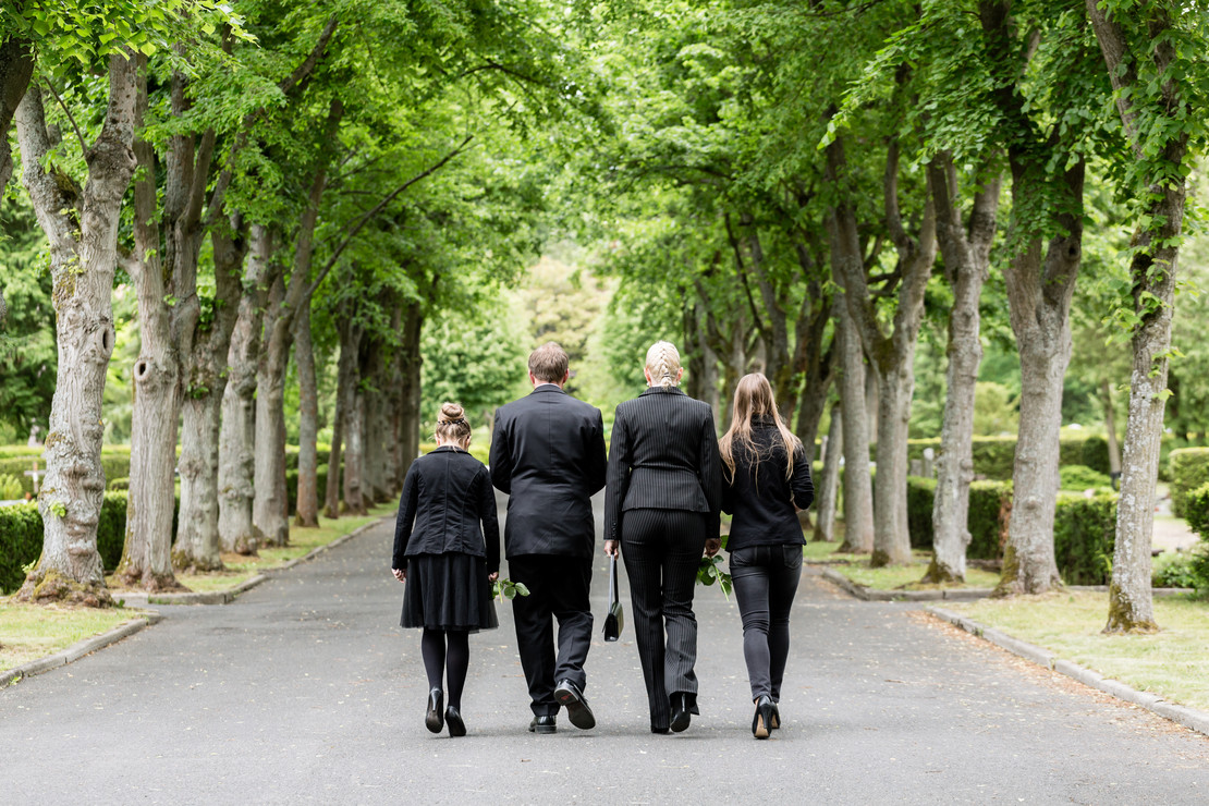 Mann, Frau und zwei Mädchen, schwarzgekleidet, von hinten, gehen eine Friedhofsallee entlang. 