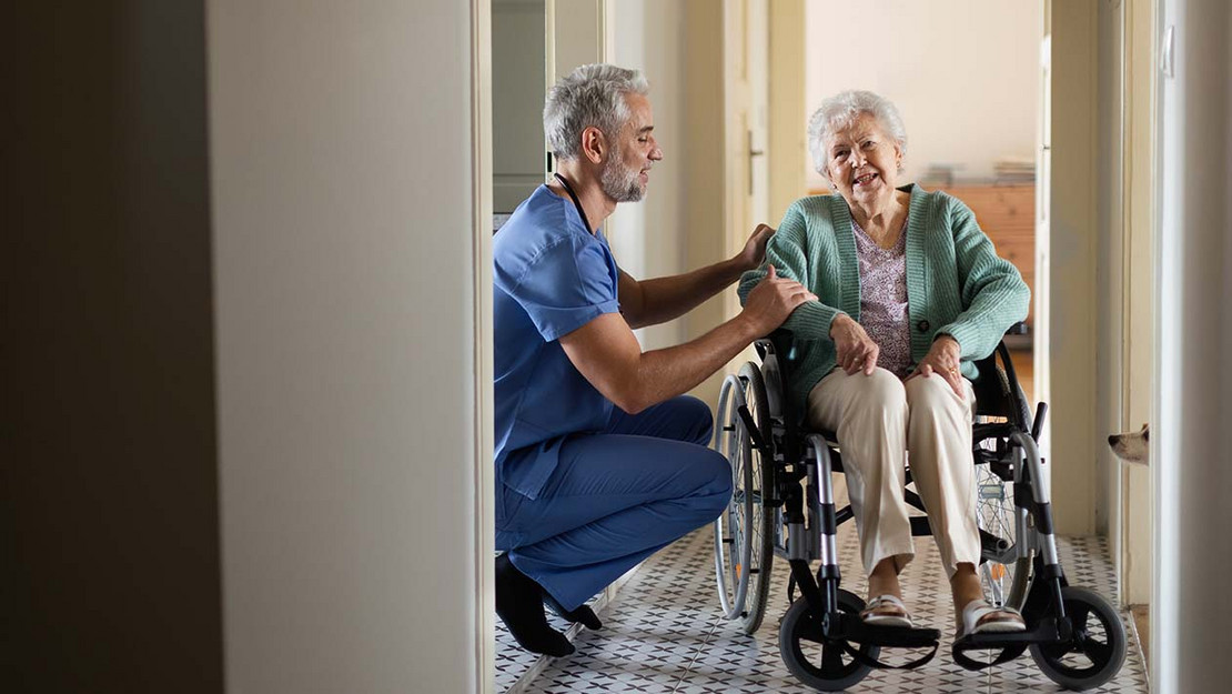 Ein Pfleger kniet neben einer Frau im Rollstuhl in deren Wohnung. 