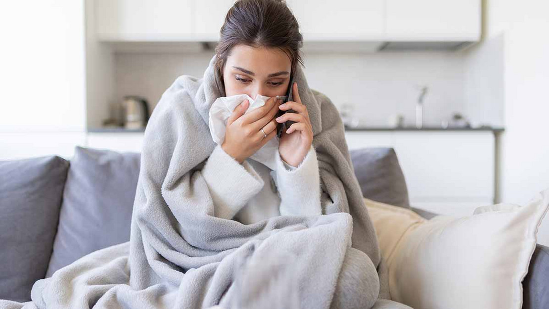 Frau sitzt in einer Decke eingewickelt auf der Couch und hält ein Taschentuch vor der Nase. Sie telefoniert. 