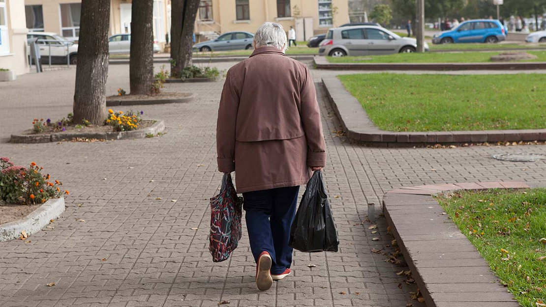 Rückenansicht einer älteren Frau, die mit zwei Beuteln durch eine urbane Landschaft geht. 
