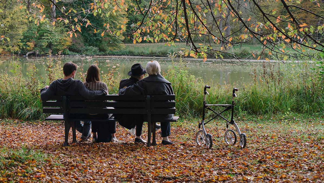 Ein junges und ein älteres Paar sitzen nebeneinander auf einer Bank an einem See in einer herbstlichen Landschaft. 