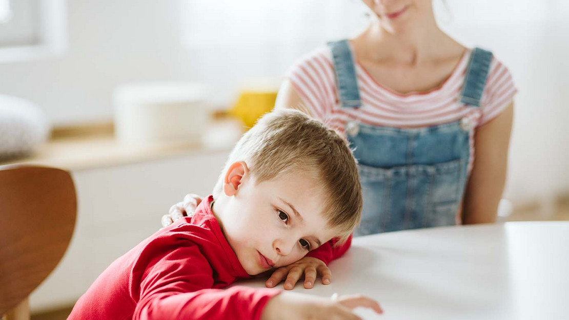 Ein Kind mit ADS oder Autismus sitzt abgelenkt an einem Schultisch, neben ihm eine Betreuerin.