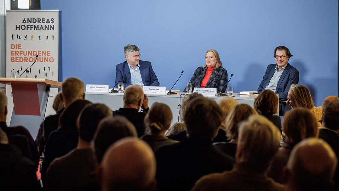 Vollbesetzter Saal, vorne ein Podium, an dem drei Personen sitzen. 